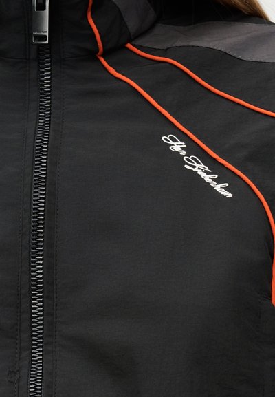 Μαύρο μπουφάν με φερμουάρ και πορτοκαλί λεπτομέρεια σε ρίγα, με λευκό κείμενο σε καλλιγραφικό στυλ "Fly Fisherman" στην περιοχή του στήθους.