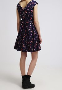 Kvinna som bär en mörkblå klänning med fjärilstryck och en rosett på ryggen, ihop med svarta ankelstövlar och strumpor, stående mot en enfärgad bakgrund.