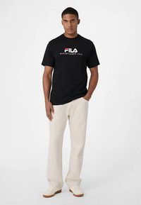 Musta lyhythihainen t-paita, jossa on rohkea valkoinen ja punainen "FILA"-logo sekä teksti "ESTABLISHED 1911", yhdistetty vaaleanbeigeihin housuihin ja valkoisiin kenkiin.