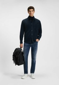 Donkerblauwe corduroy overhemd, getailleerd, met een borstzak; lichtblauwe jeans; zwarte rugzak; witte sneakers met subtiele accenten.