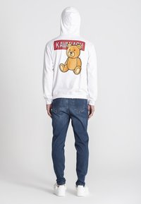 Biely mikina s veľkým grafickým znázornením medveďa a textom "KAVANACH" na chrbte, v kombinácii s tmavomodrými džínsami a bielymi teniskami.