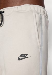 Ljusbeige Nike-mjukisbyxor med svart logotyp, utrustade med elastisk midja och en sidrand i kontrasterande ljusgrått. Mjuk tygstruktur.