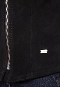 Schwarze Wildlederjacke mit einem silbernen Reißverschluss und einem kleinen metallenen Markenlabel auf der Vorderseite. Glatte Textur und figurbetontes Design.