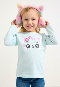 Vaaleansininen pitkähiha shirt , jossa on sarjakuvatyylinen kissan kasvot pinkillä rusetilla, yhdistetty värikkäisiin pörröisiin korvalappuihin. Lapsi hymyilee poseeratessaan.