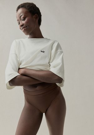 Selbstbewusste Frau mit kurzen lockigen Haaren, die ein weißes bauchfreies T-Shirt mit dem Schriftzug „self.“ und hoch taillierte braune Unterwäsche trägt, die Arme verschränkt.