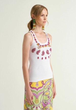 Femme aux cheveux clairs portant un haut blanc sans manches avec des motifs brodés roses et jaunes, associé à un pantalon coloré à motif cachemire et à de longues boucles d'oreilles.