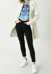 Light beige trench coat over a blue cropped sweatshirt with college print, paired with black skinny jeans and white sneakers.