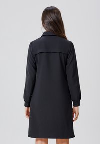 Robe noire avec un col classique, des manches longues avec des poignets élastiques, et une coupe droite qui tombe au-dessus des genoux ; texture lisse.