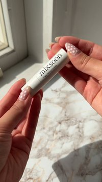 Białe cylindryczne opakowanie balsamu do ust oznaczone napisem "mixsoon" w czarnym kolorze, trzymane między palcami z jasnoróżowym wzorem paznokci z białymi kropkami.