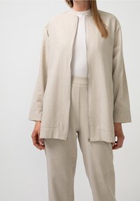 Béžová zipová bunda s kulatým výstřihem a dlouhými rukávy, vyrobená z texturované látky. V kombinaci s odpovídajícími kalhotami.
