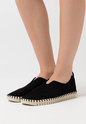 Zwarte instappers met een geperforeerde bovenkant, voorzien van een flexibele, contrasterende witte rubberen zool met ribbeldetails.