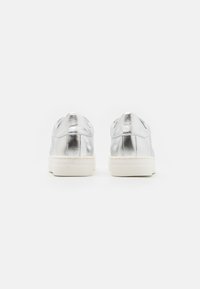 Silverfärgade metalliska sneakers med en slät yta, som har en vit gummisula och en minimalistisk design. Baksidan visar en rundad klack och sömdetaljer.