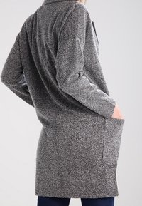 Šedý kabát s dlouhým rukávem a texturovaným vzorem, dvěma bočními kapsami a límcem. Materiál vypadá měkce a je vhodný k vrstvení.