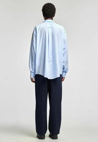 Lyseblå stripete skjorte med lange ermer, knappemansjetter og buet kant, kombinert med løstsittende marineblå bukser. Ensfarget bakgrunn.