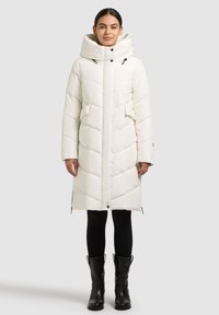 Geacă matlasată de iarnă crem, cu guler înalt, fermoar în față, model matlasat diagonal și buzunare frontale. Purtați peste colanți negri și cizme.