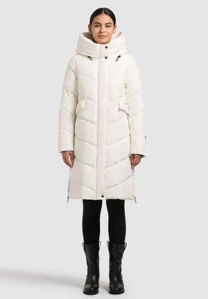 Vatteret vinterfrakke i cremefarve med høj krave, lynlåslukning foran, diagonalt quiltet mønster og forlommer. Bæres over sorte leggings og støvler.