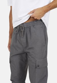 Szare spodnie cargo z elastycznym pasem i sznurkiem do regulacji. Posiada boczne kieszenie i detale z zapięciem na guzik. Tkanina jest gładka i lekka.