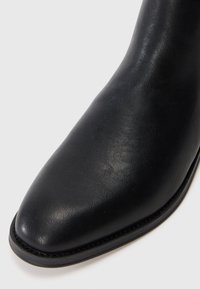 Botín negro de cuero hasta el tobillo con punta afilada, textura suave y costuras contrastantes a lo largo de la suela. Diseño clásico y acabado monocromático.