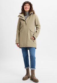 Beige parka met een capuchon, ritssluiting en drukknoopsluiting, twee voorkruiken en verstelbare manchetten, gecombineerd met blauwe jeans en bruine rubberen laarzen.