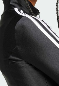 Svart sportjacka med zip, slät textur, vita ränder längs axlarna och en figursydd design som framhäver axlarna.