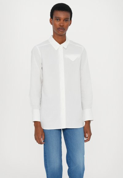 Camicia bianca a maniche lunghe con colletto button-down, dettaglio taschino frontale e una texture liscia. Abbinata a pantaloni in denim blu.