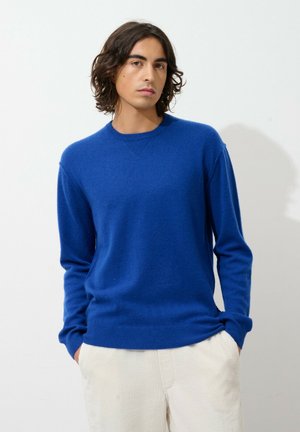 Blauwe gebreide trui met een ronde hals en lange mouwen, met ribgebreide manchetten en zoom. Draagt een crème corduroy broek.