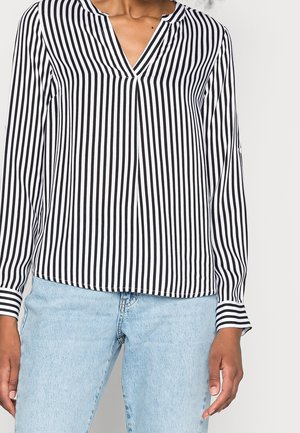 Personne portant un chemisier à rayures verticales noires et blanches à manches longues avec un col en V et un jean bleu clair.