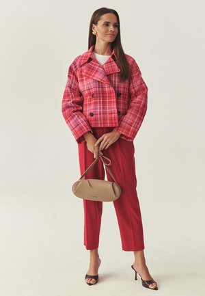 Ženska v svetlo rdečem karo suknjiču, rdečih hlačah, črnih čevljih z odprtimi prsti, ki drži bež ročno torbico z dolgim trakom na nevtralni podlagi.