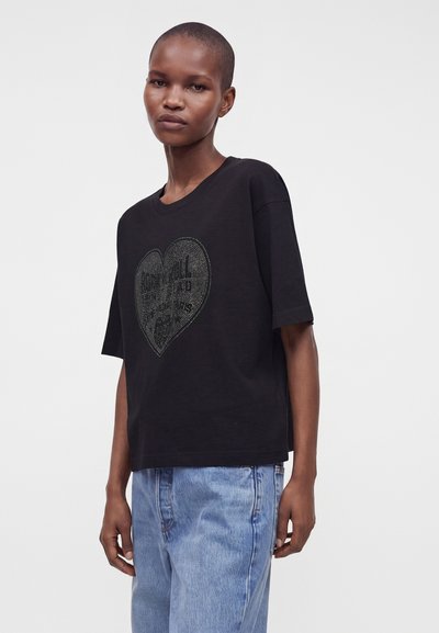Άτομο με μαύρο φαρδύ T-shirt με πουα σχέδιο καρδιάς και casual ανοιχτό μπλε τζιν, πάνω σε μονόχρωμο φόντο.