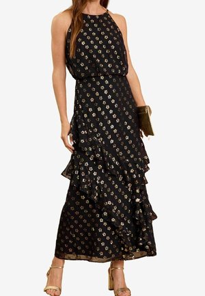 Mujer con un vestido maxi negro sin mangas con pequeño estampado floral y falda asimétrica con volantes, sosteniendo un clutch dorado y con tacones dorados.