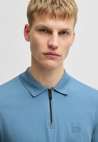 Ljusblå polo med kvartdragkedja, krag-öppning, strukturerad tyg, och en fyrkantig logotyp i en mörkare blå nyans.