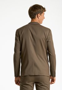 Giacca di abito marrone su misura con revers a punte, doppia spacco e spalle strutturate, caratterizzata da una consistenza liscia e un leggero luccichio.