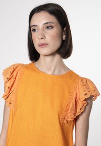 Blusa arancione con maniche a ruffle in pizzo, tessuto testurizzato, scollatura rotonda e vestibilità morbida, caratterizzata da delicati ricami sulle cuciture delle spalle.