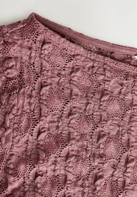 Tissu en dentelle mauve avec un motif crochet floral complexe, montrant des coutures fines et une texture délicate sur un fond clair.