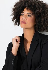 Blazer nero con revers e polsini abbottonati. La modella ha i capelli ricci e tiene la mano vicino al mento, mostrando un trucco naturale.