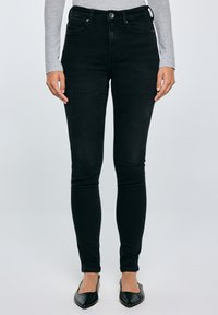 Sieviete, kas valkā augststaras melnas šauras džinsas, pelēku garām piedurknēm krekliņu un melnas punktveida kurpes, stāvot pret vienkrāsainu fonu.