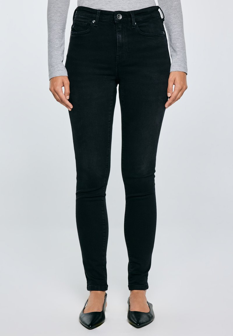 Sieviete, kas valkā augststaras melnas šauras džinsas, pelēku garām piedurknēm krekliņu un melnas punktveida kurpes, stāvot pret vienkrāsainu fonu.