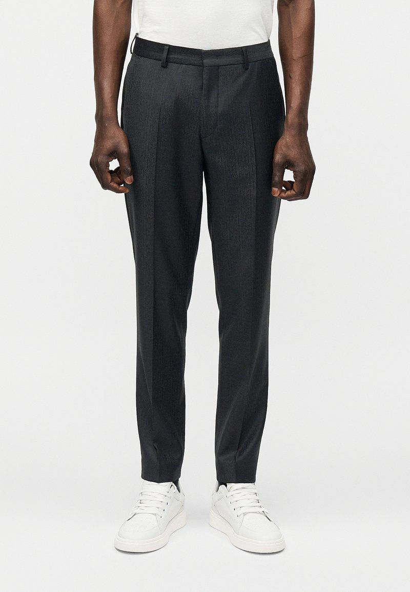 Homme portant un pantalon noir sur mesure avec des plis devant et des baskets blanches, debout devant un fond uni.