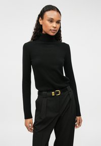 Fekete garbó pulóver puha anyagból, hosszú ujjal és testhezálló fazonnal. Magas derekú fekete nadrággal és arany csatú övvel viselve.