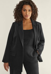Blazer nero con vestibilità sartoriale, chiusura a un bottone, revers a scollo, e tasche frontali; abbinato a un top nero. Tessuto dalla texture liscia.