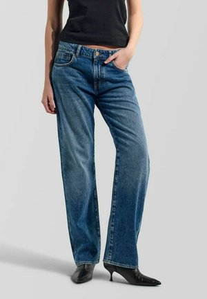 Personne portant un jean bleu large à taille haute et des chaussures à talon kitten noires à bout pointu, debout avec une main dans la poche.