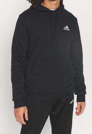 Mann trägt schwarzen Adidas-Hoodie und passende schwarze Adidas-Hose mit weißen Logos, steht vor einfarbigem Hintergrund.