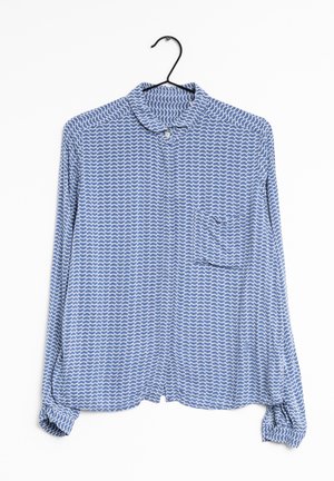 Sininen pitkähihainen kauluspaita valkoisella aaltokuvioinnilla, roikkumassa mustassa henkari valkoista taustaa vasten.