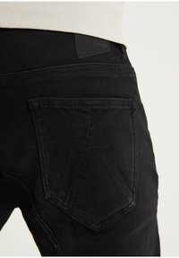 Los jeans de mezclilla negra presentan una tela texturizada, diseñados con una forma de bolsillo única y un parche de cuero negro en la cintura.