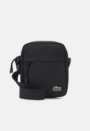 Petit sac bandoulière noir compact avec sangle réglable, poche avant zippée et petit logo crocodile vert en bas à droite.