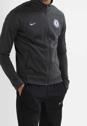 Person wearing a black zip-up sports jacket with Chelsea FC logo and matching black Nike track pants, hands in pockets.