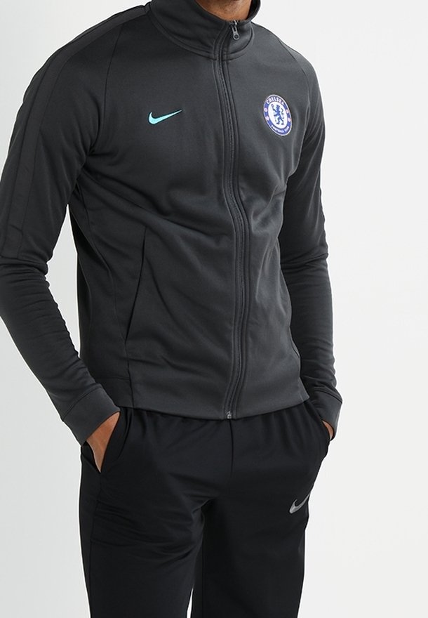 Personne portant une veste de sport noire zippée avec le logo du Chelsea FC et un pantalon de survêtement noir assorti de la marque Nike, mains dans les poches.