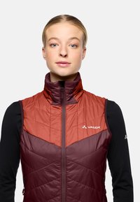 Rood en bordeaux gekleurde geïsoleerde vest met een hoge kraag, ritsluiting en een chevronpatroon. Voorzien van een logo op de bovenste linkerzijde en een gladde textuur.