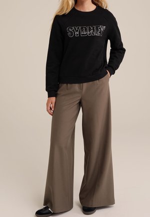 Vrouw die een zwart sweatshirt met de tekst "SYDNEY" en wijde bruine broek draagt, poseert met één hand in haar zak tegen een effen achtergrond.