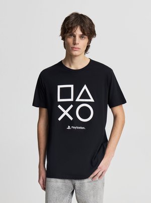 Giovane uomo che indossa una t-shirt PlayStation nera con simboli bianchi quadrato, triangolo, X e cerchio, abbinata a jeans grigio chiaro.